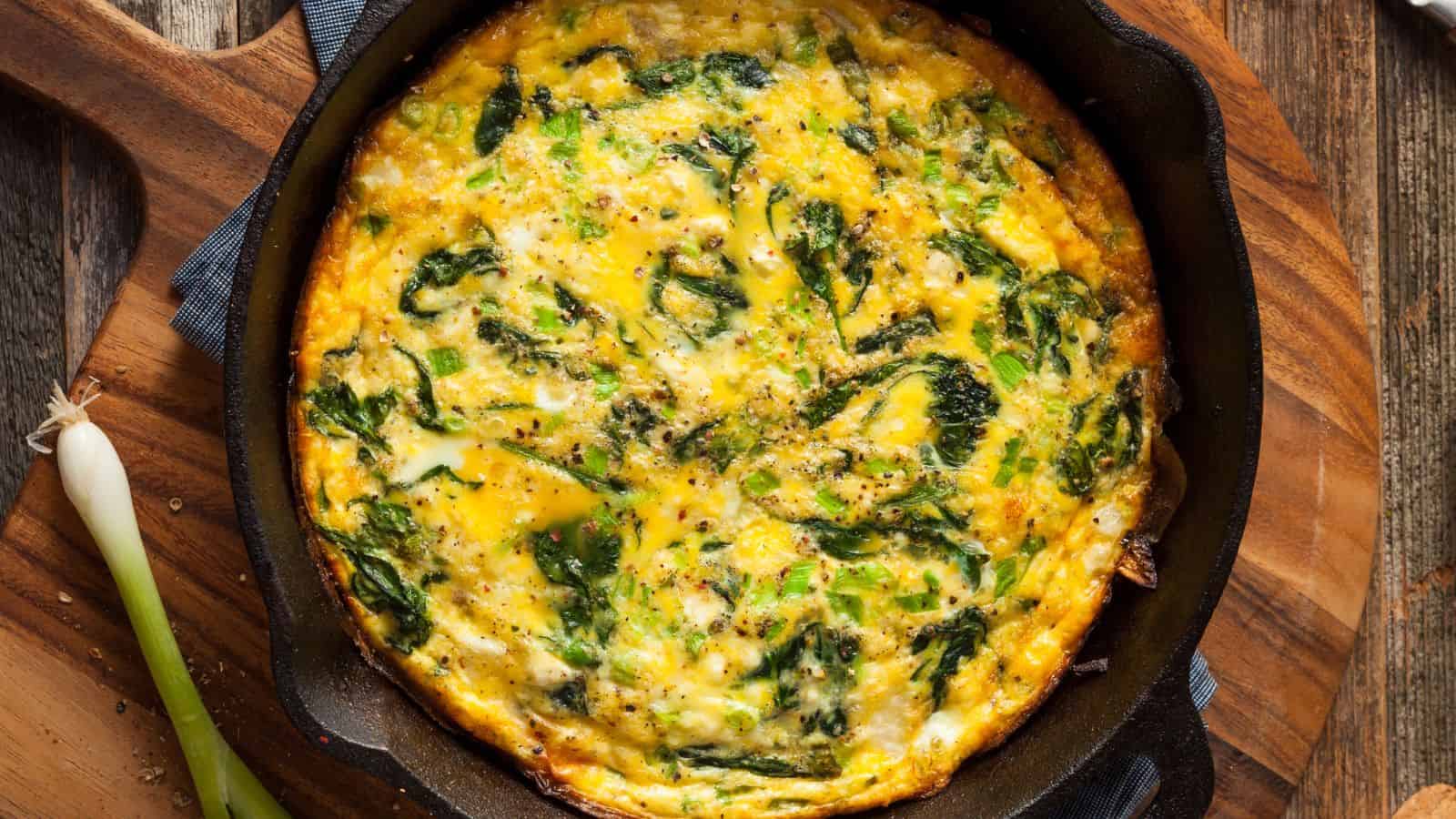 A baked spinach and cheese frittata, perfect for high-protein breakfasts, sits in a black cast iron skillet on a wooden surface, with a green onion nearby.