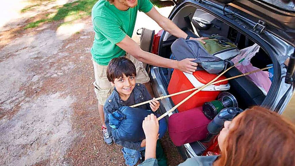A family loads camping gear, including a cooler and sleeping bags, into a car's trunk. A young boy holds a sleeping bag and smiles at the camera.