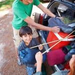 A family loads camping gear, including a cooler and sleeping bags, into a car's trunk. A young boy holds a sleeping bag and smiles at the camera.