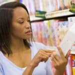 A woman stands in a grocery store aisle, reading the information on the side of a boxed product while surrounded by shelves of various food items—perhaps checking to see if it complies with California’s new food law.