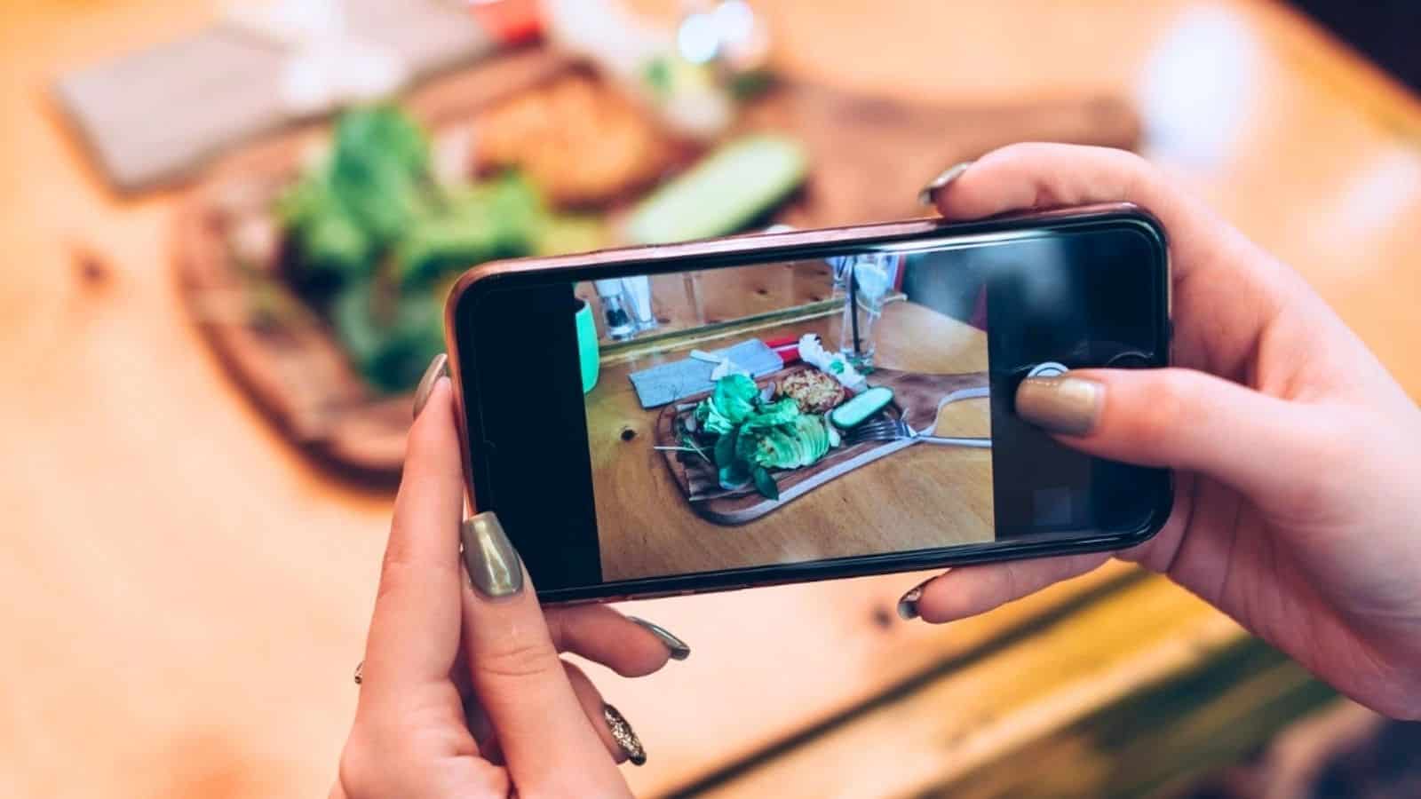 A person holds a smartphone and takes a photo of a plate of food on a wooden table.