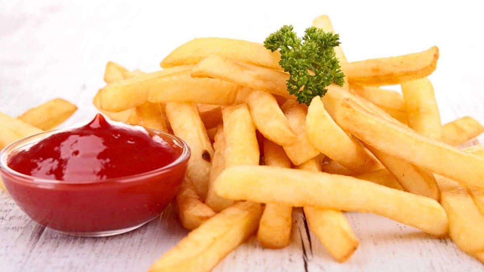 A pile of French fries garnished with parsley next to a small glass bowl of ketchup on a white surface.