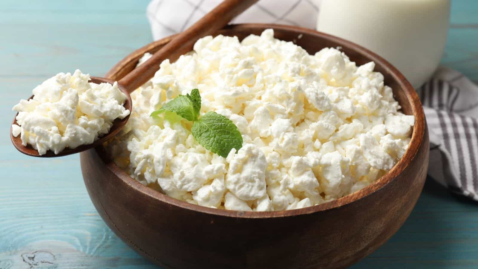 A wooden bowl filled with creamy cottage cheese, garnished with a fresh mint leaf, sits on a blue table next to a spoon and a striped cloth.