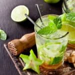 Two refreshing mojito cocktails with lime slices, mint leaves, and ice are on a wooden board, surrounded by fresh lime, starfruit, mint, brown sugar cubes, and a wooden muddler—perfect for crafting the ultimate mojito experience.
