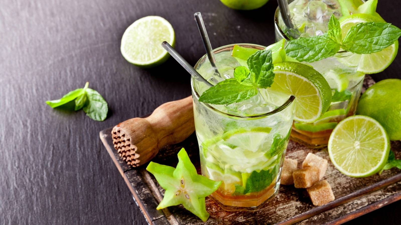 Two refreshing mojito cocktails with lime slices, mint leaves, and ice are on a wooden board, surrounded by fresh lime, starfruit, mint, brown sugar cubes, and a wooden muddler&mdash;perfect for crafting the ultimate mojito experience.