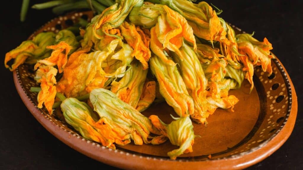 A brown ceramic plate holds fresh squash blossoms with yellow and orange petals, displayed against a dark background.