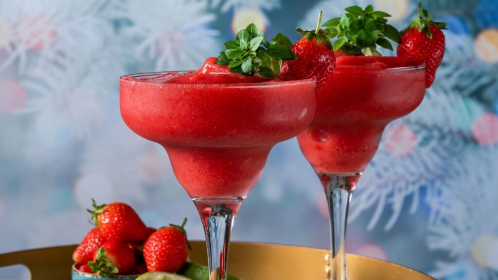 Two strawberry margaritas in wide glasses, garnished with fresh strawberries and mint, are displayed on a tray with whole strawberries and limes nearby. Reminiscent of a refreshing daiquiri, this vibrant scene is set against frosted greenery in the blurred background.