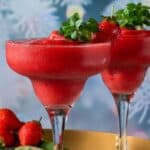 Two strawberry margaritas in wide glasses, garnished with fresh strawberries and mint, are displayed on a tray with whole strawberries and limes nearby. Reminiscent of a refreshing daiquiri, this vibrant scene is set against frosted greenery in the blurred background.