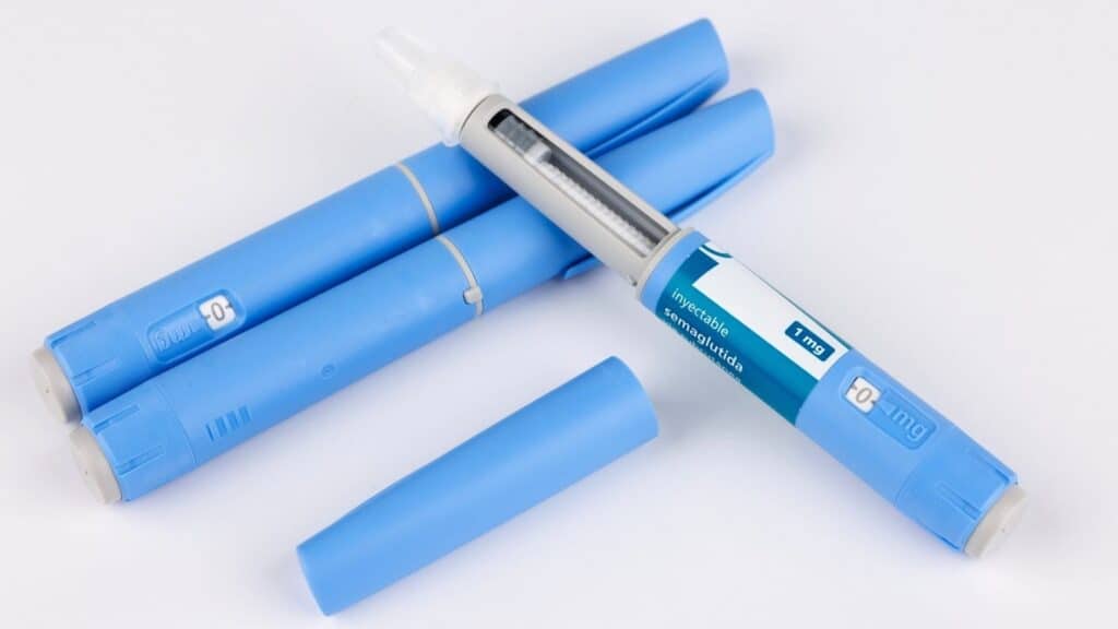 Four blue insulin pens are shown, one uncapped with a visible needle and dosage label, and three capped, arranged on a white background. These pens are similar in appearance to some GLP-1 drugs used for diabetes management.