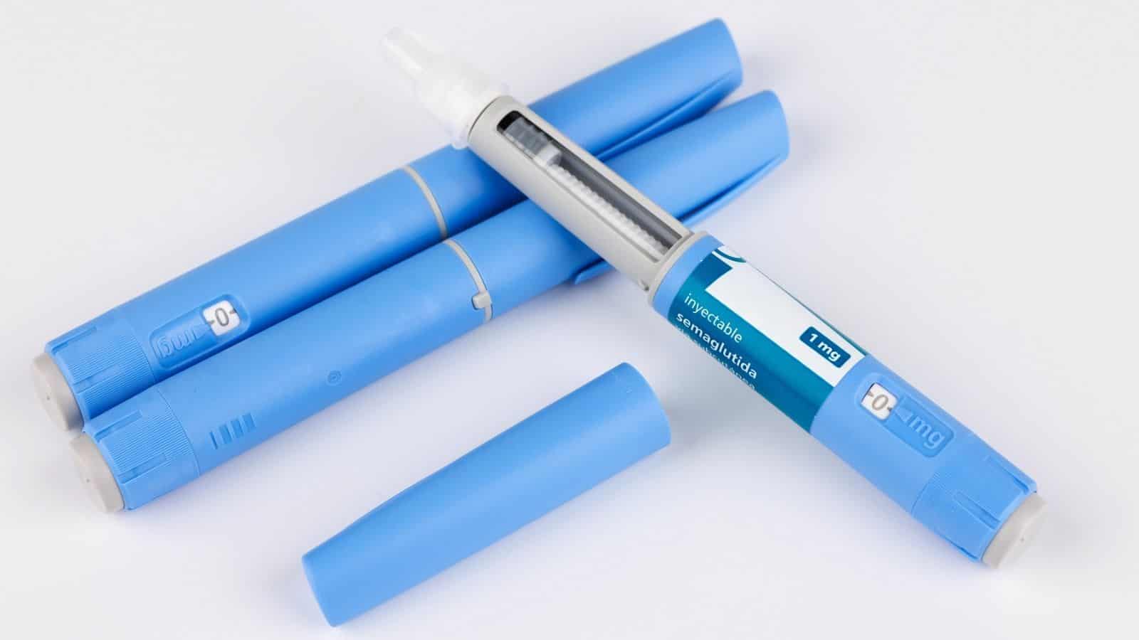 Four blue insulin pens are shown, one uncapped with a visible needle and dosage label, and three capped, arranged on a white background. These pens are similar in appearance to some GLP-1 drugs used for diabetes management.