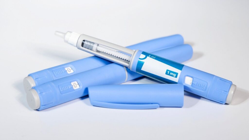 Three blue medical injection pens are shown, one with the cap off revealing the needle and dosage window labeled "1 mg.