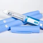 Three blue medical injection pens are shown, one with the cap off revealing the needle and dosage window labeled "1 mg.