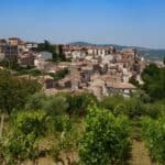A hillside village with stone houses and red roofs sits above a vineyard and olive trees under a clear blue sky, evoking the timeless charm of southern Italy wine estates.