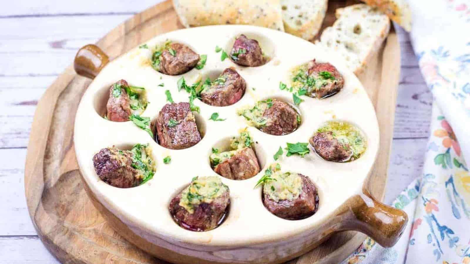 Garlic Butter Steak Bites Escargot-Style dish in a round ceramic plate with cooked snails topped with herbs and butter sauce, and slices of bread on a wooden board.