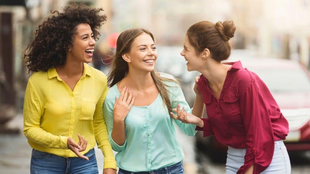 Three young women smiling and talking while walking on a city street, wearing colorful shirts.