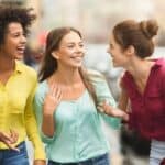 Three young women smiling and talking while walking on a city street, wearing colorful shirts.