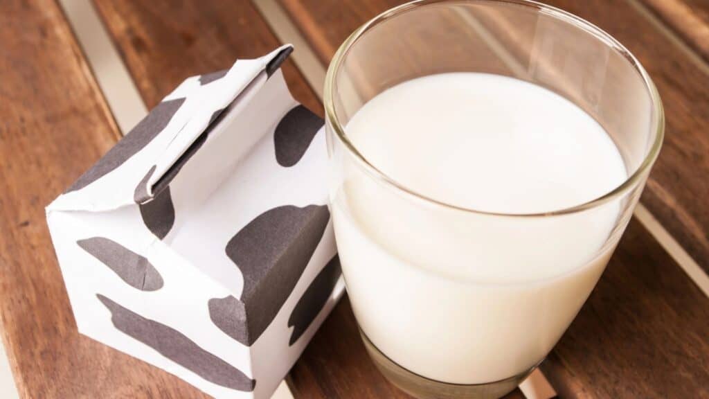 A glass of milk sits next to a small, cow-patterned milk carton on a wooden table.