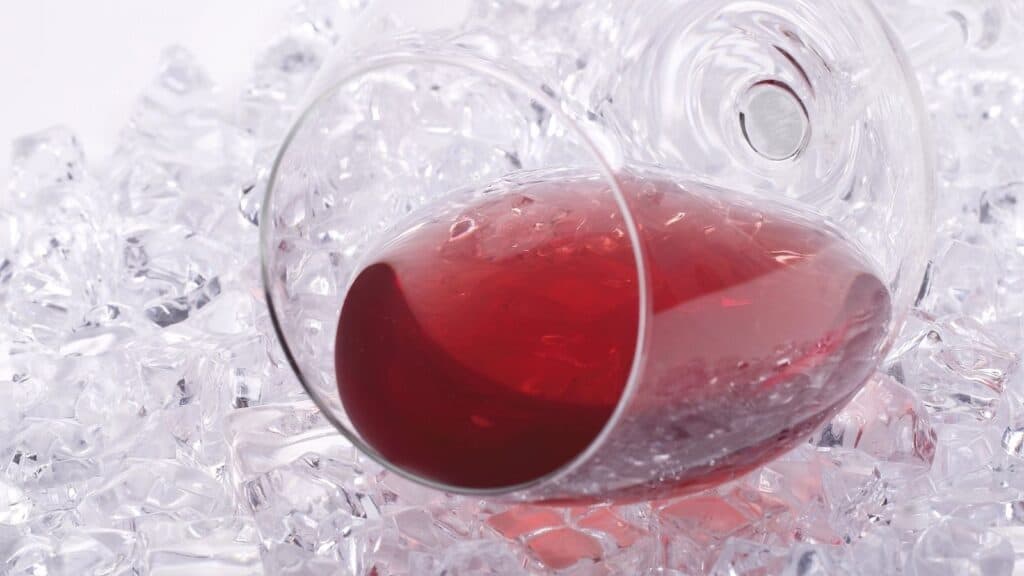 A wine glass filled with red liquid lies on its side atop a bed of clear ice cubes.