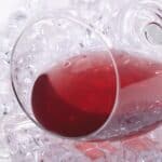 A wine glass filled with red liquid lies on its side atop a bed of clear ice cubes.