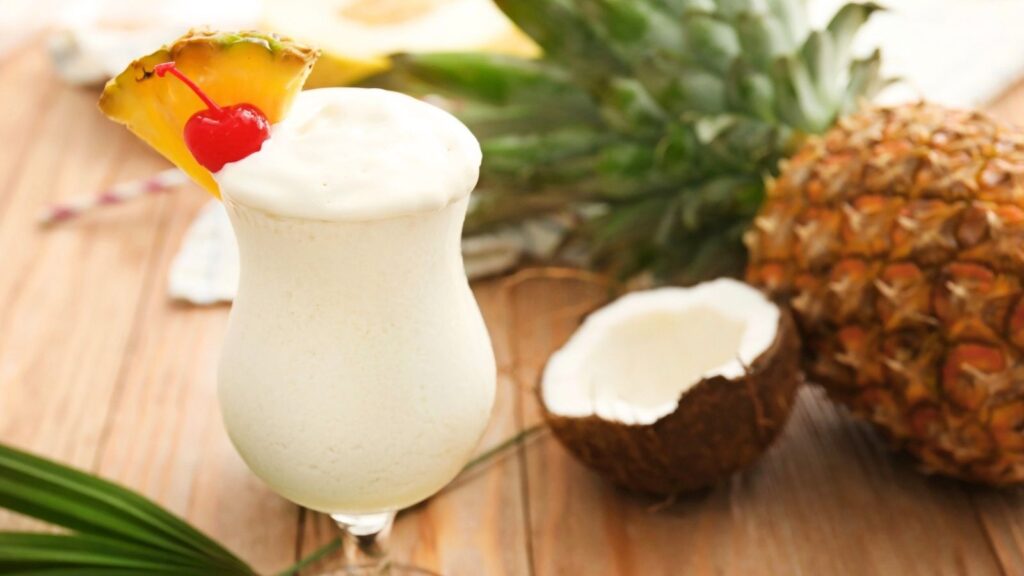 A pi&ntilde;a colada made with premium rum, garnished with a pineapple slice and cherry, sits on a table next to a whole pineapple and a halved coconut.