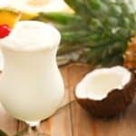 A piña colada made with premium rum, garnished with a pineapple slice and cherry, sits on a table next to a whole pineapple and a halved coconut.