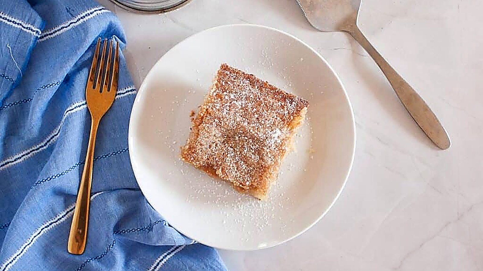 A square piece of Gooey Butter Cake. topped with powdered sugar on a white plate, next to a gold fork, a blue cloth, and a metal cake server on a marble surface.