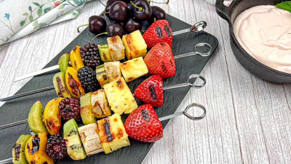 Four metal skewers with grilled fruit and cheese cubes are arranged on a black tray, accompanied by fresh cherries and a bowl of creamy dip.