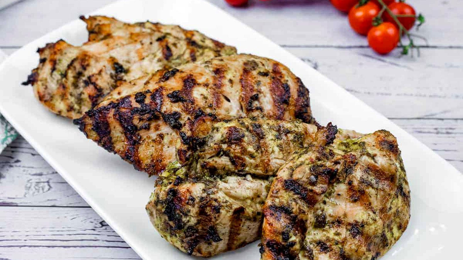 Grilled chicken on a wooden board with cilantro and tomatoes in the back.
