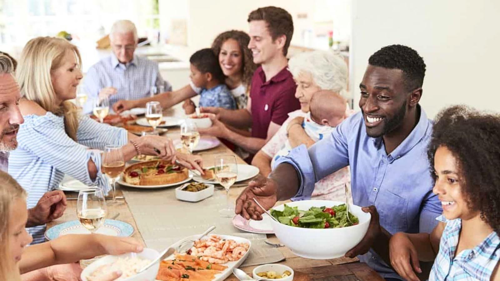 A group of people of various ages sit around a dining table, sharing a meal with pizza, salad, shrimp, and wine in a bright, homey setting.