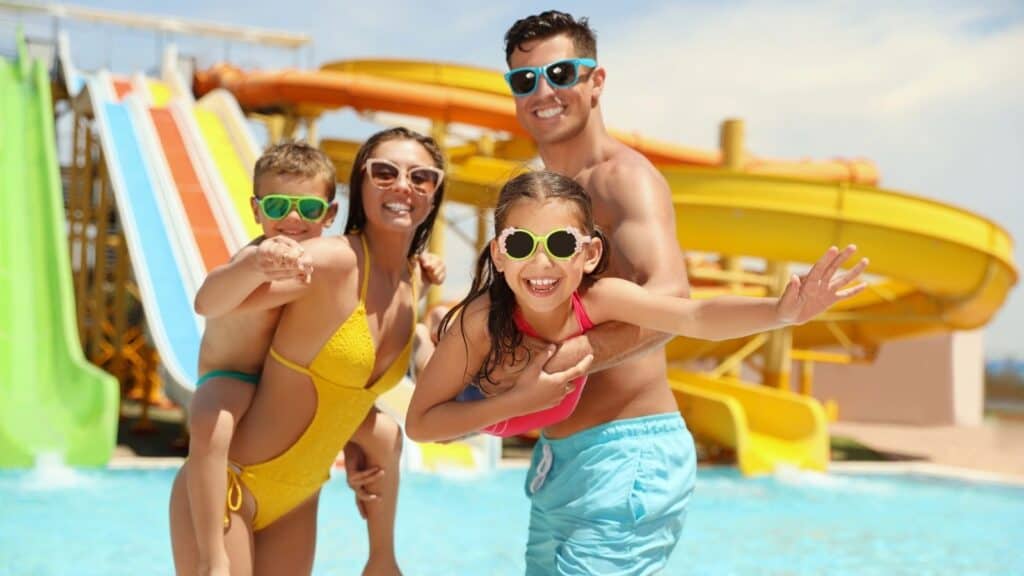 Four people in swimsuits and sunglasses stand by water slides at a water park, smiling at the camera as they celebrate National Waterpark Day.