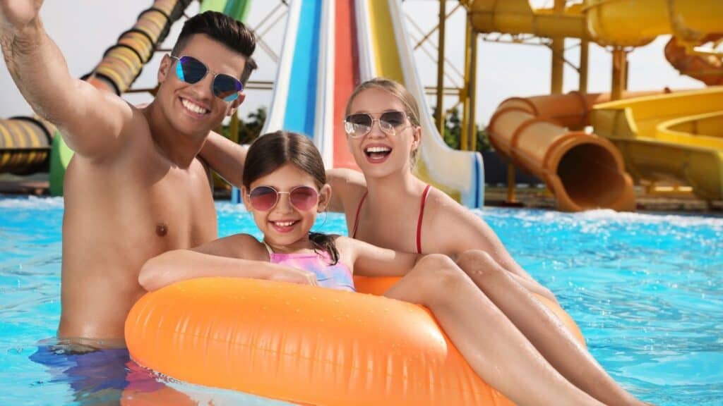 Three people wearing sunglasses smile while sitting on an orange pool float in a lively waterpark pool, with colorful slides visible in the background.