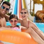 Three people wearing sunglasses smile while sitting on an orange pool float in a lively waterpark pool, with colorful slides visible in the background.