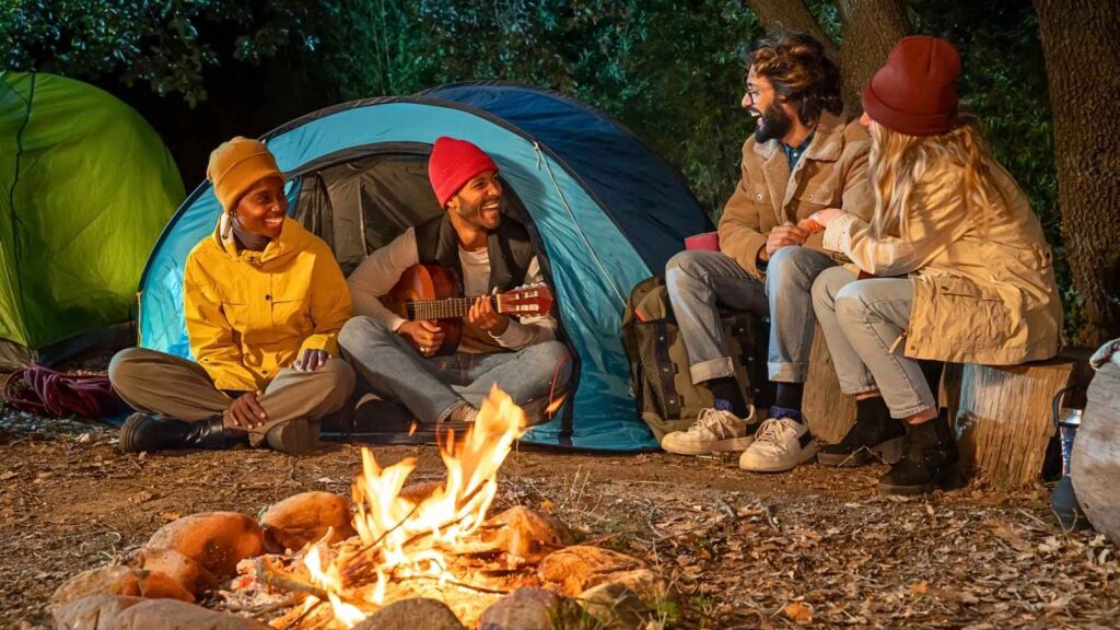 Four people enjoy a screen-free camping experience as they sit around a campfire near a blue tent at night; one person plays guitar while the others listen and talk, all wearing jackets and hats.