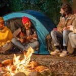 Four people enjoy a screen-free camping experience as they sit around a campfire near a blue tent at night; one person plays guitar while the others listen and talk, all wearing jackets and hats.