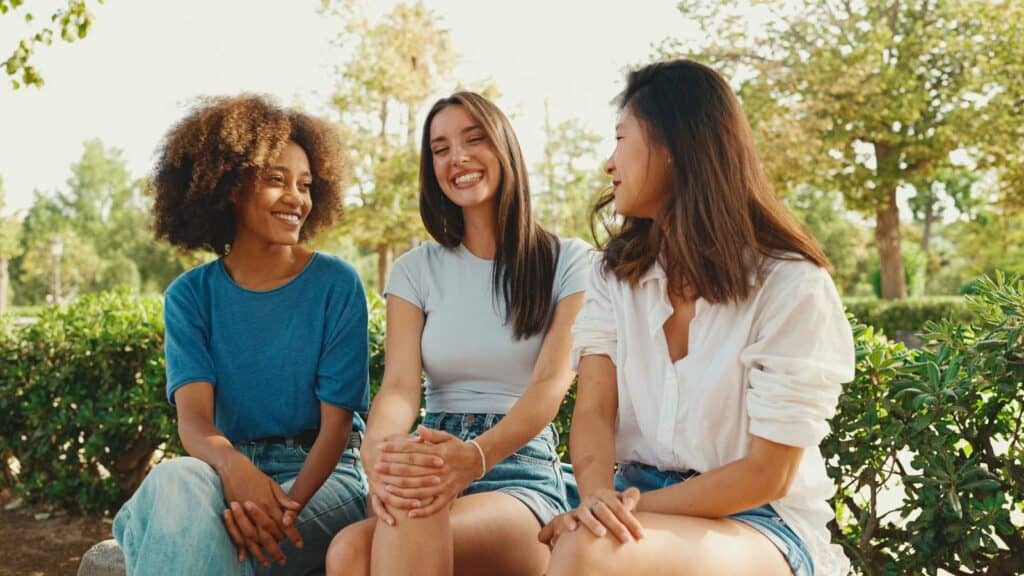 Three young women sit on a bench outdoors, smiling and talking with trees and greenery in the background.