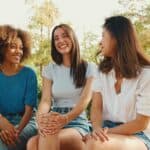 Three young women sit on a bench outdoors, smiling and talking with trees and greenery in the background.