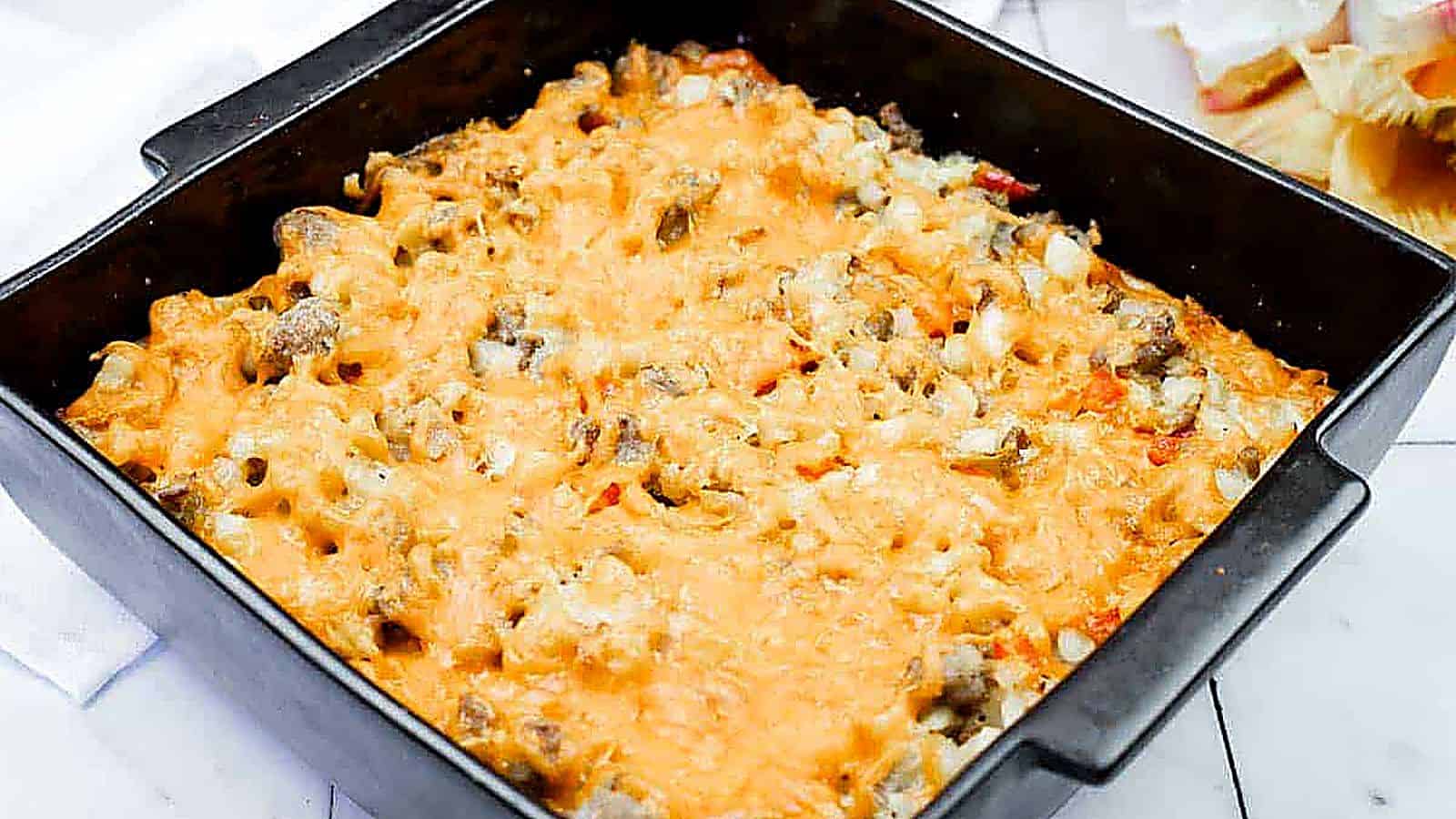 A square black baking dish filled with a baked casserole topped with melted cheese, containing visible pieces of meat and vegetables.