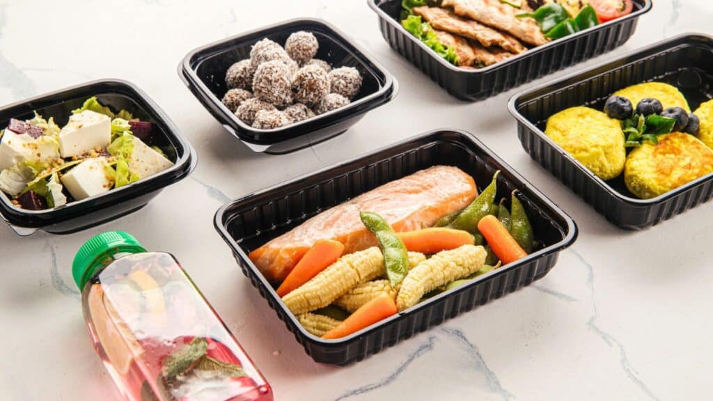 Assorted meal prep containers with salmon and vegetables, salad with feta, meatballs, omelet patties with berries, grilled chicken salad, and a bottle of infused water on a white surface.