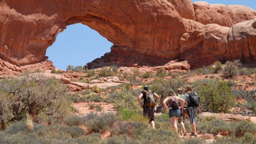 Three people with backpacks walk toward a large natural rock arch in a desert landscape with sparse vegetation and a clear blue sky.