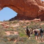 Three people with backpacks walk toward a large natural rock arch in a desert landscape with sparse vegetation and a clear blue sky.