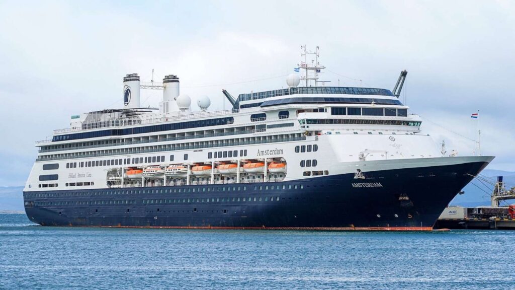 A large cruise ship with multiple decks is docked at a port. The ship is white and navy blue with lifeboats visible along the side.