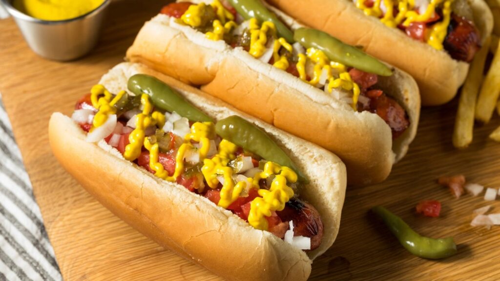Three hot dogs topped with chopped onions, relish, mustard, peppers, and ketchup are arranged on a wooden board with a side of French fries.