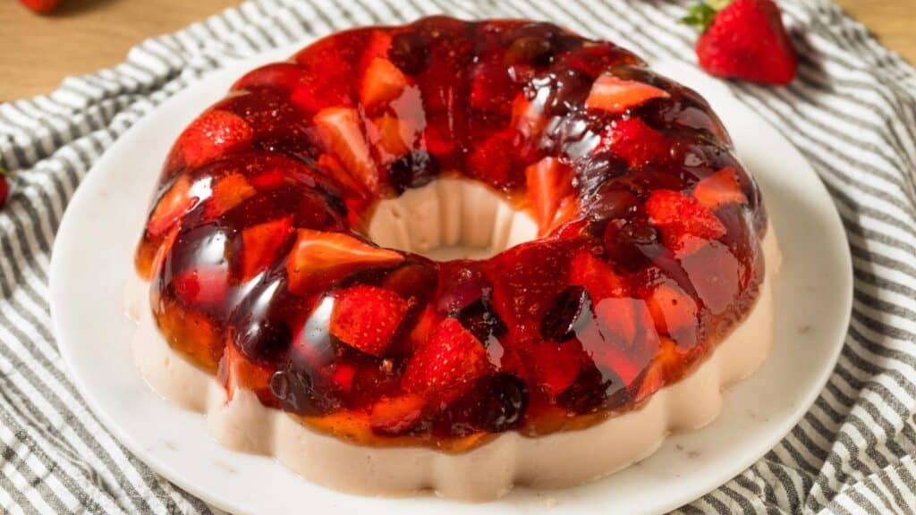 A gelatin dessert ring with visible strawberries and fruit pieces, served on a white plate atop a striped cloth.