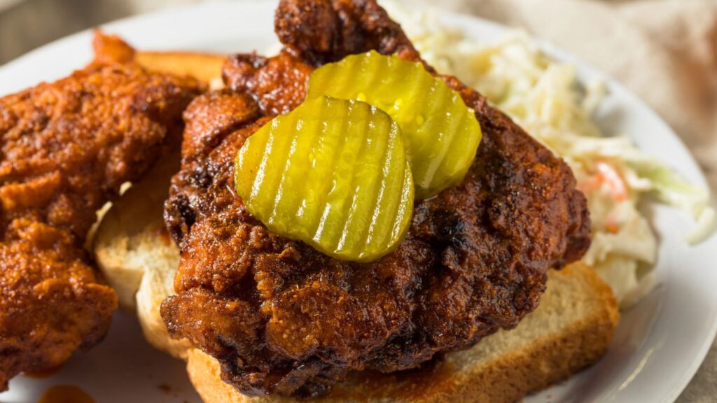 A plate with fried chicken on sliced bread, topped with pickle chips, with coleslaw on the side.