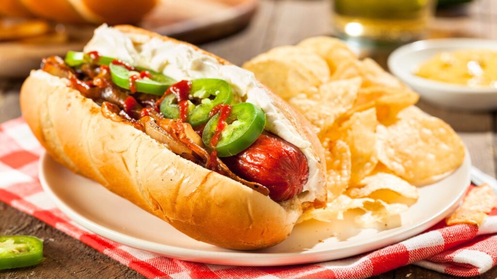 A hot dog topped with sliced jalapeños, grilled onions, ketchup, and mayonnaise, served with potato chips on a white plate.