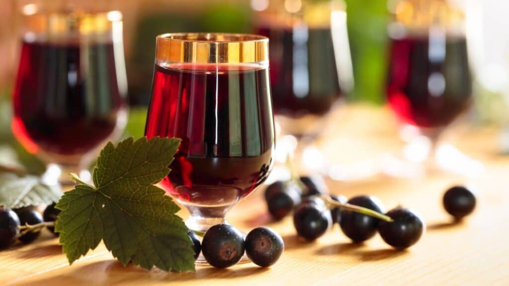 Four glasses of dark red liquid on a table, surrounded by blackcurrants and green leaves, with sunlight in the background.