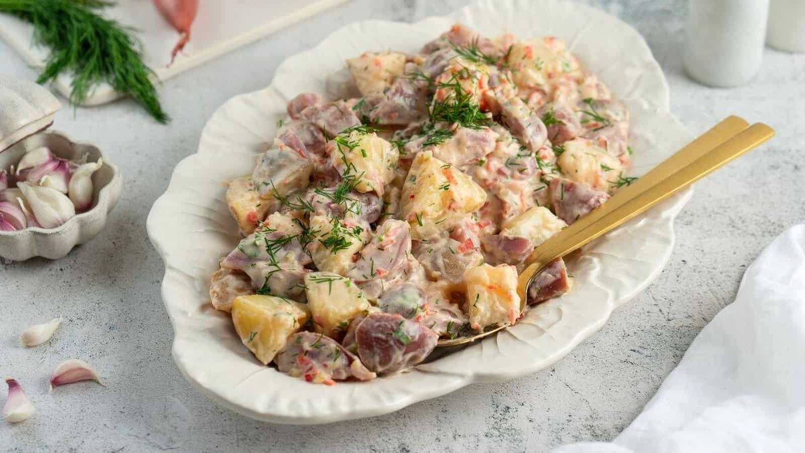 A serving dish filled with Classic Red Potato Salad with Pickles is garnished with fresh dill, with a gold-colored serving spoon resting in the dish.