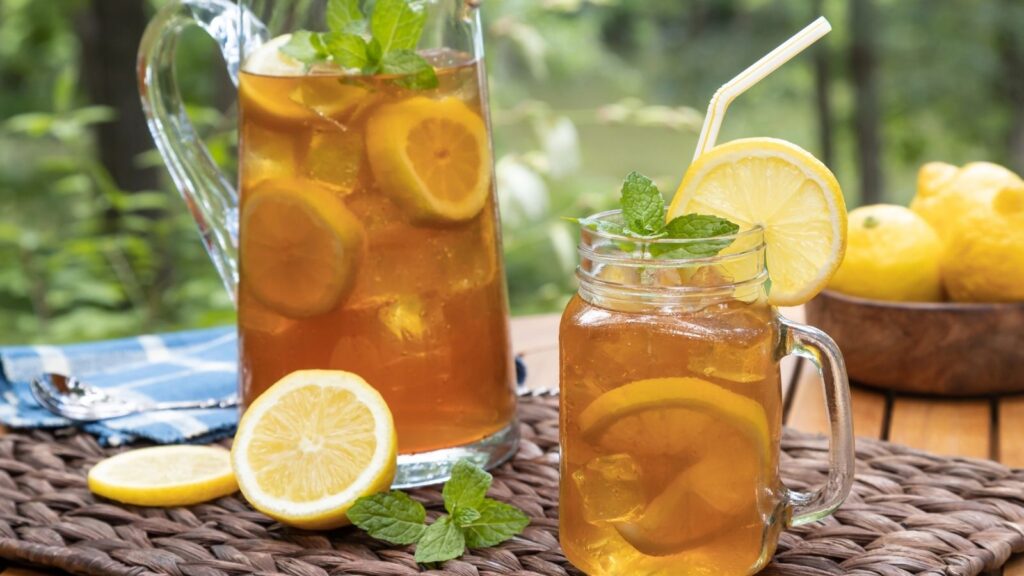 A glass and pitcher of iced tea with lemon slices, mint leaves, and ice cubes sit on a woven mat next to fresh lemons, reminiscent of the refreshing experience you get with McAlister's free tea.