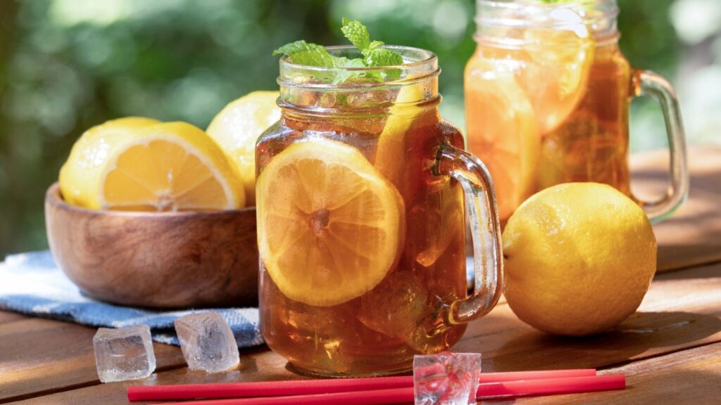 Two mason jars of iced tea with lemon slices and mint on a wooden table, next to whole lemons, a bowl of lemons, ice cubes, and red straws&mdash;perfect for enjoying McAlister's free tea on a warm day.