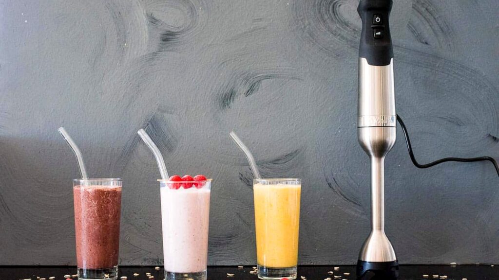 Three smoothies in glasses with straws—chocolate, pink with raspberries, and yellow—are lined up next to an immersion blender against a gray background.